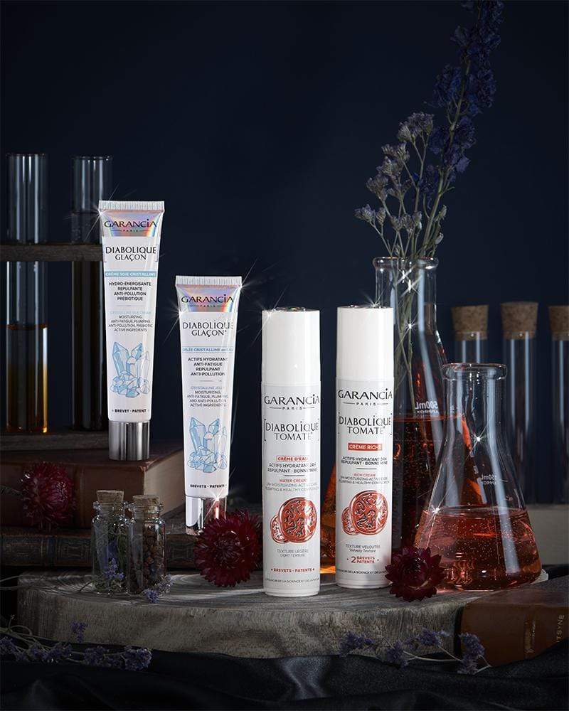 Laboratoire Garancia Crème de jour DIABOLIQUE GLAÇON CRÈME SOIE CRISTALLINE : Mise en scène de produits cosmétiques Garancia dans une ambiance d'apothicaire mystique, incluant les gammes Diabolique Glaçon et Diabolique Tomate entourées de fioles de laboratoire, de fleurs séchées et de vieux livres.