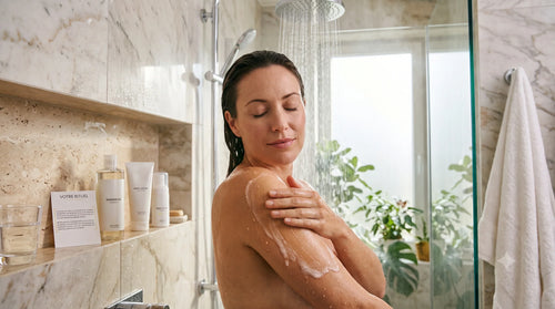 Une femme se lave sereinement sous la douche en massant son bras avec une huile lavante qui se transforme en fine mousse, à côté de flacons de soin neutres posés sur une étagère en marbre.