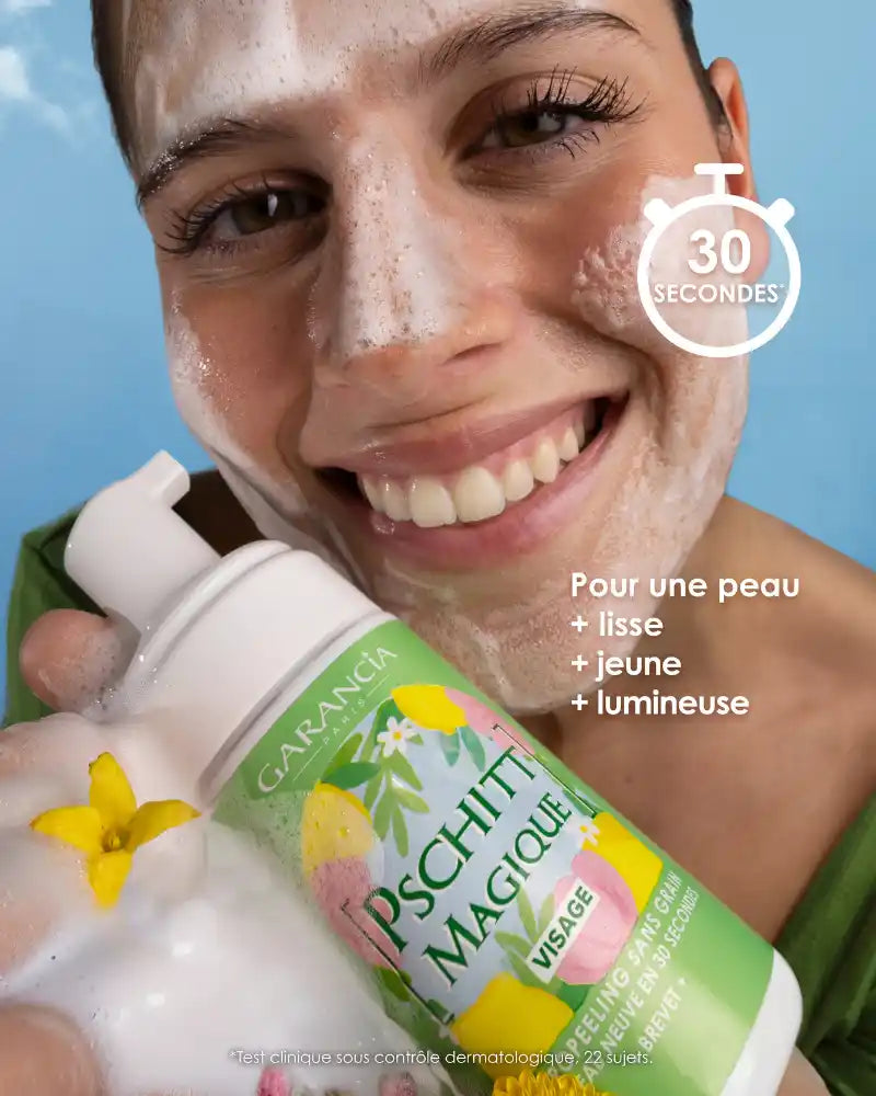 Une femme souriante applique la mousse onctueuse du Pschitt Magique Garancia sur son visage. Elle tient le flacon édition limitée devant un ciel bleu parsemé de nuages, illustrant un moment de soin frais et naturel.