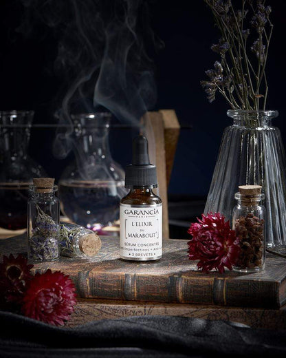 Sérum L'Élixir du Marabout de Garancia posé sur un vieux grimoire, entouré de fleurs rouges, de fioles d'apothicaire et d'une fumée mystique évoquant un laboratoire de sorcellerie.